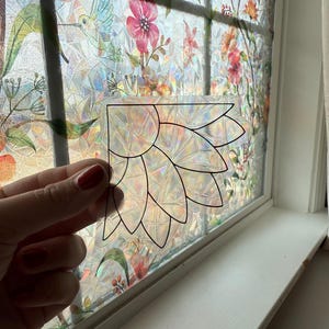 May include: A clear, decorative window film with a floral design is held in front of a window with a similar pattern. The film features a black outline of a stylized flower. The window and film have a rainbow-like, iridescent effect.