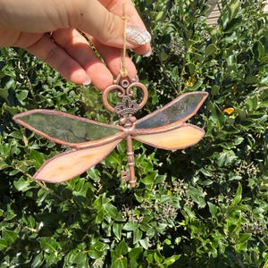 Puede incluir: Adorno de libélula de vidriera con cuerpo en forma de llave y detalles de corona. Las alas son una mezcla de vidrio verde y melocotón, con un marco de metal color cobre. Se adjunta una cuerda dorada para colgar.