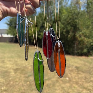 May include: A collection of colorful stained glass surfboard pendants, each with a silver-toned metal outline. The surfboards are in shades of green, blue, red, orange, and yellow, suspended on tan cords. The pendants are held up against a blurred outdoor background.