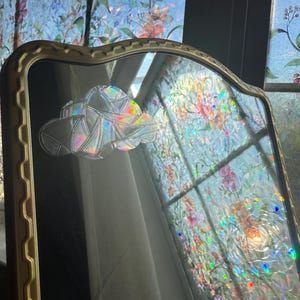 May include: A decorative mirror with a gold-colored frame. The mirror's surface features a cloud-shaped design with iridescent, rainbow-colored accents. The background shows a window with floral patterned stained glass.