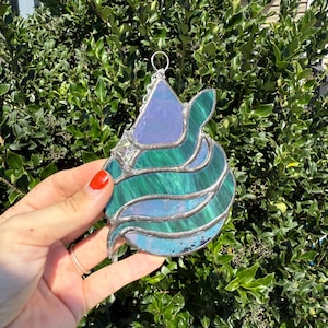 May include: A stained glass ornament in shades of blue and green, with a silver-colored metal border. The ornament has a unique shape, with a triangular top and a swirling, layered design. It is hanging in front of a green bush.