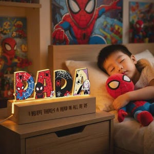 以下が含まれることがあります： スパイダーマンをテーマにした文字でDADAと綴られた木製のライトアップサイン。赤、青、黒の色合いで、ベースには「I BELIEVE THERE'S A HERO IN ALL OF US」というテキストが刻印されており、内部の光源によって照らされています。