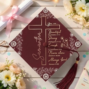 Op de afbeelding: Een bordeauxrode afstudeerhoed met een gouden kruis en de naam "Samantha". De hoed heeft inspirerende tekst en bloemmotieven. Een bordeauxrode kwast hangt aan de hoed. De afbeelding bevat een diploma, bloemen en een notitieboek.