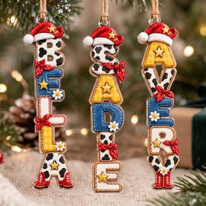 Puede incluir: Tres adornos colgantes verticales con los nombres BELLA, BEADIE y AVERY. Cada letra está decorada con estampados de vaca, cuadros amarillos o texturas azules, acentuadas con botas de vaquero rojas, pañuelos rojos y estrellas doradas.