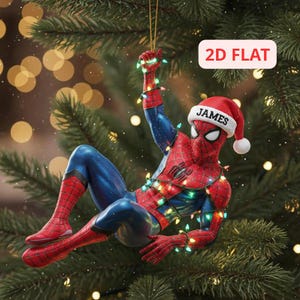 Puede incluir: Un adorno de árbol de Navidad de Spider-Man que muestra al personaje con un traje rojo y azul, usando un gorro de Papá Noel con el nombre JAMES impreso en el borde blanco. La figura está envuelta en luces navideñas de colores.