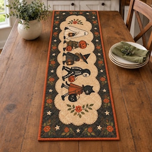 Peut inclure: Un chemin de table matelassé rectangulaire avec un panneau central crème à bordure festonnée. Le motif représente cinq personnages en costumes d'Halloween, dont un fantôme, une citrouille, une sorcière, un squelette et un chat noir, tenant des seaux.