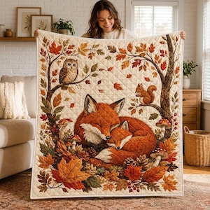 May include: A quilted throw blanket featuring a printed scene of two sleeping foxes nestled among autumn leaves, pinecones, and mushrooms. The design includes a tree with an owl perched on a branch and a squirrel, all set against a cream background.