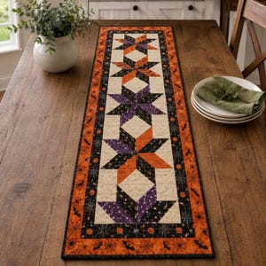May include: A rectangular quilted table runner featuring a central row of four star-shaped blocks in orange, purple, and black fabric. The border displays a pattern of orange pumpkins, black bats, and white spiderwebs on a black background.