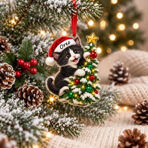 Puede incluir: Un adorno navide&ntilde;o colgante que muestra a un gatito blanco y negro con un gorro de Pap&aacute; Noel rojo con el nombre Oreo impreso en el borde. El gatito abraza un peque&ntilde;o &aacute;rbol de Navidad cubierto de nieve y decorado con bolas rojas y verdes.