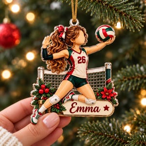 Puede incluir: Un adorno decorativo que muestra a una jugadora de voleibol con cabello largo castaño, vistiendo una camiseta blanca y verde con el número 26, sosteniendo un balón. El diseño incluye una red, bayas de acebo y una placa con el texto Emma.