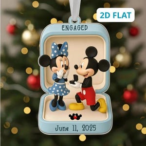 Puede incluir: Un adorno en forma de caja de anillo azul claro con Mickey y Minnie Mouse. Mickey está arrodillado sosteniendo un anillo, mientras Minnie lleva un vestido azul de lunares y un lazo. El texto dice ENGAGED y June 11, 2025, con un corazón.