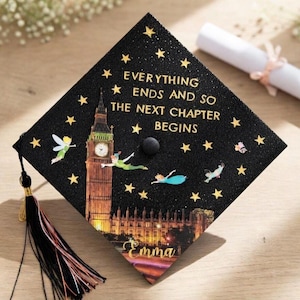 May include: Black graduation cap with gold text reading "EVERYTHING ENDS AND SO THE NEXT CHAPTER BEGINS." Features a Big Ben illustration, Peter Pan characters, and gold stars. Includes a black tassel and the name "Emma".