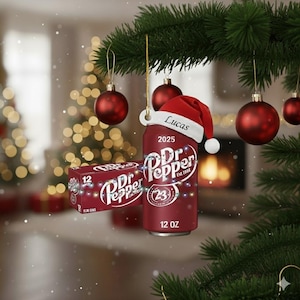 Puede incluir: Adorno navideño con forma de lata de Dr. Pepper, adornado con un gorro de Papá Noel con el nombre "Lucas" y el año "2025". El adorno cuelga de una rama de árbol de Navidad, acompañado de bolas rojas y una caja de latas de Dr. Pepper. La lata dice "12 OZ".