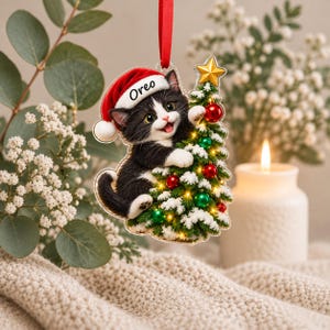Puede incluir: Un adorno navide&ntilde;o colgante que muestra un gatito blanco y negro con un gorro de Pap&aacute; Noel rojo con el nombre Oreo impreso en el borde. El gatito trepa por un peque&ntilde;o &aacute;rbol de Navidad verde cubierto de nieve y decorado con bolas rojas y verdes.