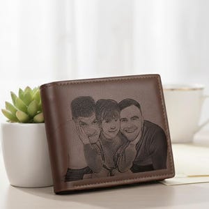 Puede incluir: Cartera de cuero marrón con una imagen personalizada de tres personas. La cartera rectangular presenta un retrato grabado detallado. Una planta suculenta en una maceta blanca y una taza blanca están en el fondo.