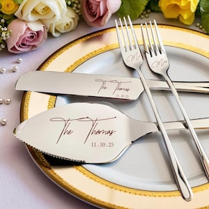 May include: Silver wedding cake serving set with forks, a knife, and a server. The set is engraved with "The Thomas" and the date 11.10.25. The forks have an "M" engraved on them. The set is on a gold-rimmed plate.