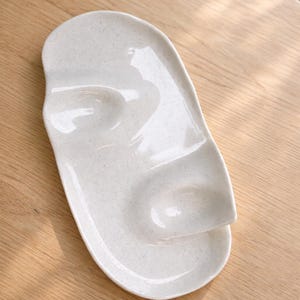 May include: A speckled, off-white ceramic serving platter with a unique face-like design. The platter features two recessed areas and a raised, curved section resembling a nose. The oval-shaped dish is suitable for serving appetizers or small dishes.