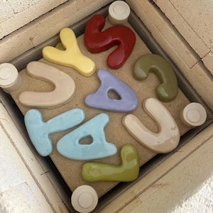 May include: A selection of brightly coloured ceramic letters inside a kiln. The letters are in pastel shades such as yellow, red, blue, and green. They are arranged on a brown surface, with small white ceramic cylinders around the edges.
