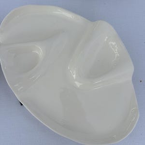 May include: A white ceramic serving dish with three compartments. The dish has an unusual, organic shape with smooth, curved edges. The glossy surface reflects light, highlighting its clean design. The dish is likely used for serving appetizers or snacks.