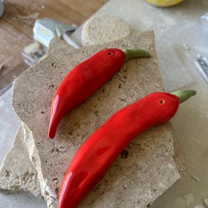 May include: Two glossy red ceramic chilli peppers with green stalks are displayed on a textured stone surface. The peppers have a smooth, curved shape and a vibrant colour. The image suggests a kitchen or home decor theme.