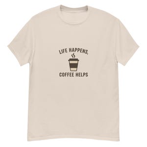 May include: A light beige t-shirt with the text "LIFE HAPPENS, COFFEE HELPS" in brown. A brown coffee cup graphic is centered between the text. The shirt has short sleeves and a crew neck.