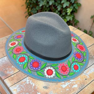 Puede incluir: Un sombrero fedora gris de ala ancha con un patrón floral pintado a mano. El diseño incluye flores vibrantes en rojo, rosa, morado y blanco con hojas verdes y líneas onduladas azules que rodean la copa sobre la superficie del ala.