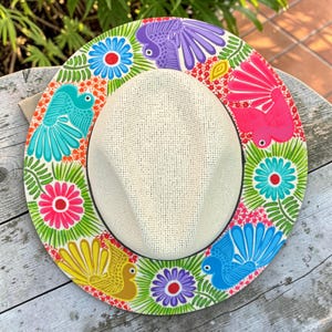 Puede incluir: Un sombrero de paja trenzada de color crema con un ala ancha pintada a mano con un patrón vibrante y colorido. El diseño incluye aves estilizadas en morado, rosa, turquesa y amarillo, intercaladas con motivos florales brillantes y follaje verde.