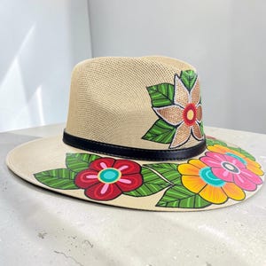 Puede incluir: Un sombrero fedora de paja beige con una banda de cuero negro y diseños florales pintados a mano. La copa muestra una flor con hojas verdes, mientras que el ala ancha está decorada con un grupo de flores vibrantes en rojo, amarillo y rosa.