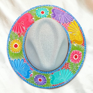 Puede incluir: Un sombrero fedora de color gris claro con un ala ancha pintada a mano con un vibrante patrón de flores y aves. El diseño incluye pájaros rosas, morados, amarillos y azules intercalados con flores coloridas y hojas verdes sobre un fondo gris.