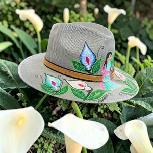 Puede incluir: Un sombrero fedora de color gris claro con una banda de cuero marrón y diseños pintados a mano de calas azules y blancas con hojas verdes. Una pequeña figura con un atuendo turquesa y rosa está pintada en el lateral de la copa.