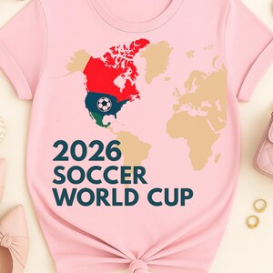 May include: Pink t-shirt featuring a world map design with a focus on North America, colored in red, green, and blue. The text "2026 SOCCER WORLD CUP" is printed in dark teal. A soccer ball graphic is included.