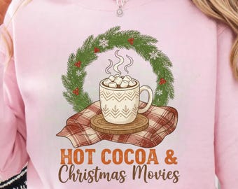 May include: A light pink sweatshirt featuring a graphic of a steaming mug of hot cocoa with marshmallows, set against a green wreath with red berries. The text "HOT COCOA & Christmas Movies" is below the graphic.