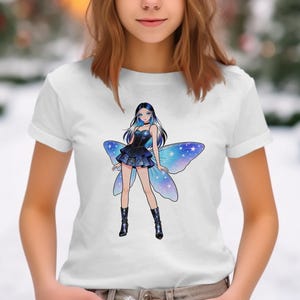 May include: White t-shirt featuring a cartoon illustration of a fairy with blue and black hair, a dark blue dress, and blue butterfly wings. The fairy is wearing black boots. The background is blurred.