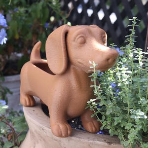 May include: A brown dachshund-shaped planter, perfect for a garden or patio. The planter is filled with soil and surrounded by green plants and blue flowers. The dog planter is made of a durable material.