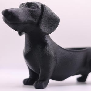 May include: A black dachshund figurine, crafted with a textured finish. The dog is depicted in a standing pose, with its head tilted upwards. The figurine has a small opening on its back, suggesting it could be a planter or a decorative item.