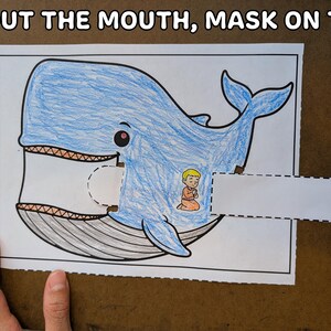 May include: A blue whale coloring page with a cut-out mouth and a small figure inside. The text "6. OUT THE MOUTH, MASK ON TOP" is at the top. The whale is drawn with blue crayon and has a detailed mouth.