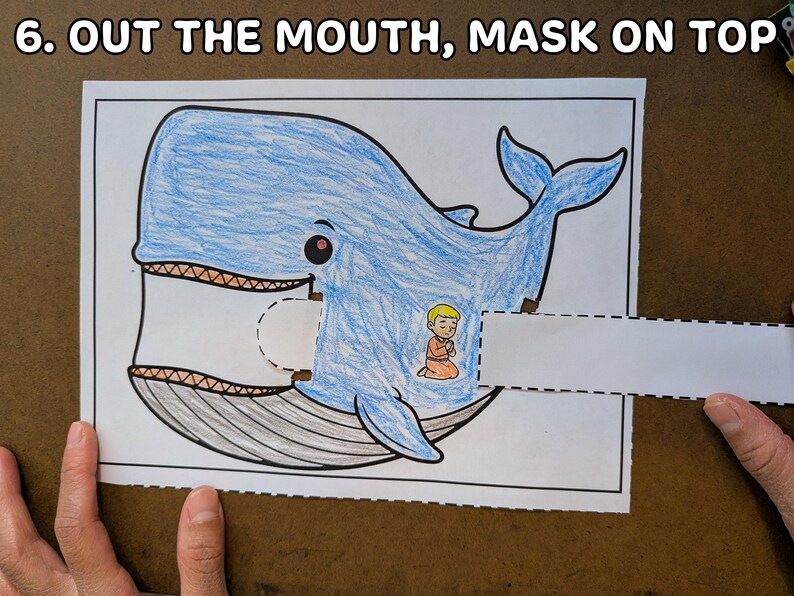 May include: A blue whale coloring page with a cut-out mouth and a small figure inside. The text "6. OUT THE MOUTH, MASK ON TOP" is at the top. The whale is drawn with blue crayon and has a detailed mouth.