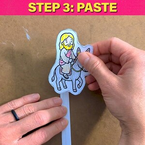 May include: A colorful illustration of a religious figure riding a donkey is being pasted onto a white strip. The image is part of a craft project, with the text "STEP 3: PASTE" in yellow on a pink background.