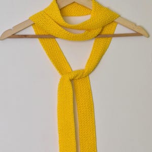 May include: A vibrant yellow knitted scarf hangs on a wooden clothes hanger against a white backdrop. The scarf is knotted at the bottom, with the ends falling downwards. The texture of the knit is clearly visible, indicating a handcrafted item.