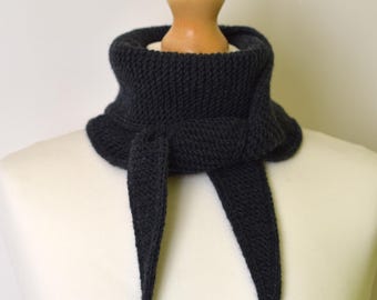 May include: A dark charcoal grey knitted neck warmer featuring a textured garter stitch pattern. The accessory is designed with a wide, folded collar section that wraps around the neck and secures with two pointed, dangling ends tied in a knot.