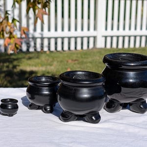 Pu&ograve; includere: Una collezione di quattro vasi in ceramica nera lucida di varie dimensioni, ciascuno con un corpo arrotondato e tre piedini. I vasi sono disposti su una superficie bianca, con uno sfondo sfocato di una recinzione bianca e erba verde.