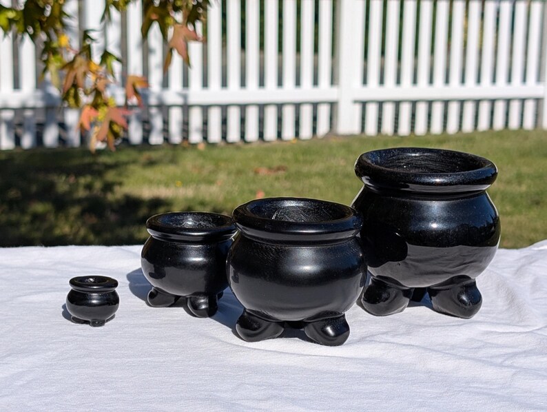 Pu&ograve; includere: Una collezione di quattro vasi in ceramica nera lucida di varie dimensioni, ciascuno con un corpo arrotondato e tre piedini. I vasi sono disposti su una superficie bianca, con uno sfondo sfocato di una recinzione bianca e erba verde.