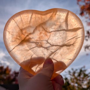 May include: A translucent, heart-shaped stone with a warm, peachy-orange hue. The stone has natural veining and cracks throughout, catching the light. The heart is held up against a backdrop of a blue sky and autumn foliage.