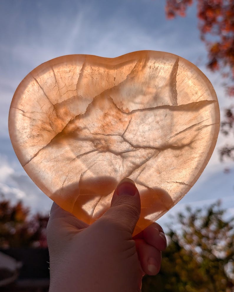 May include: A translucent, heart-shaped stone with a warm, peachy-orange hue. The stone has natural veining and cracks throughout, catching the light. The heart is held up against a backdrop of a blue sky and autumn foliage.
