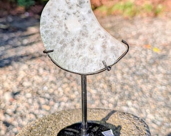 May include: A crescent moon-shaped slice of translucent white quartz crystal with a textured, crystalline surface. The stone is held by a black metal wire cradle mounted on a thin vertical rod and a circular black metal base stand.