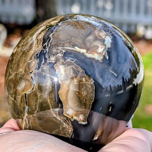 May include: A polished, spherical stone with a marbled appearance. The orb features a mix of brown, tan, and black hues, with intricate patterns and natural veining. The surface is glossy and reflects light, creating a captivating display.