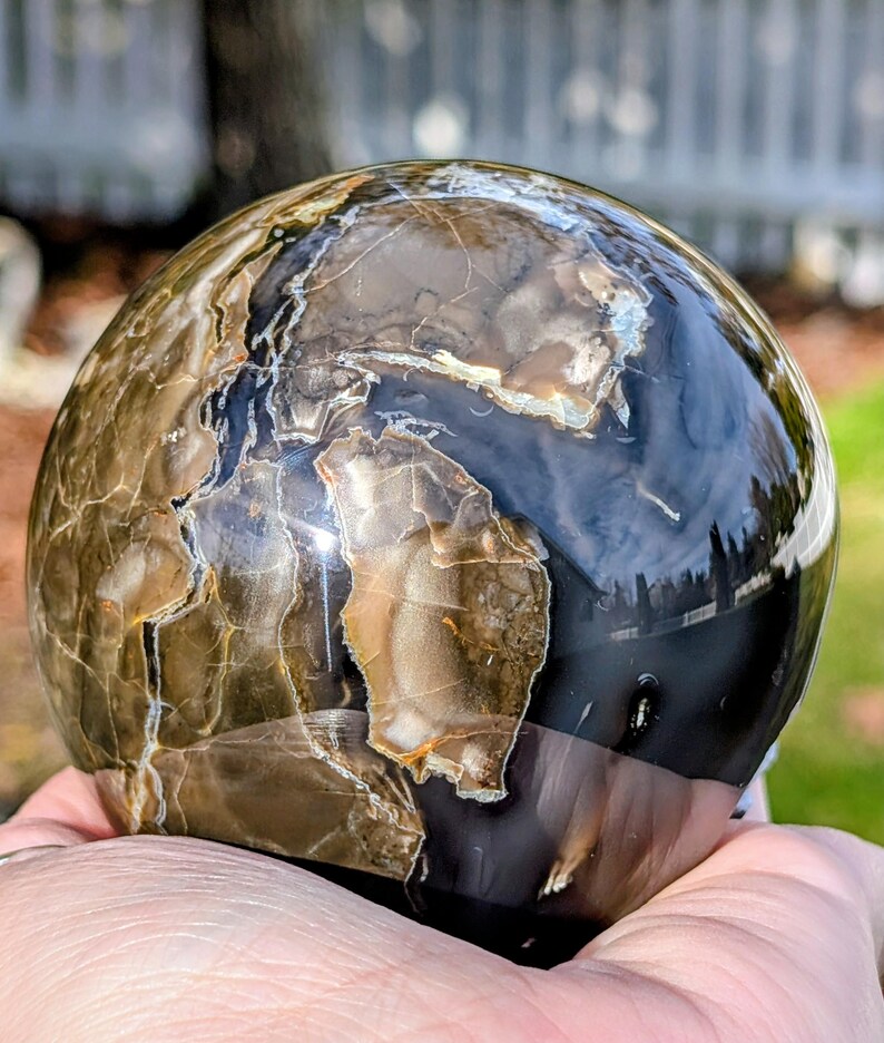 May include: A polished, spherical stone with a marbled appearance. The orb features a mix of brown, tan, and black hues, with intricate patterns and natural veining. The surface is glossy and reflects light, creating a captivating display.