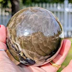 May include: A polished, spherical gemstone with a marbled pattern in shades of brown, gray, and cream. The sphere is held in a hand, reflecting the surrounding environment. The surface has natural veining.