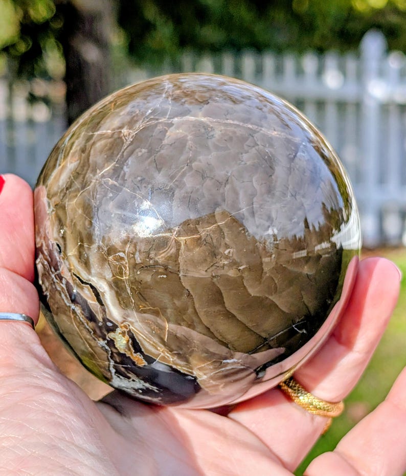 May include: A polished, spherical gemstone with a marbled pattern in shades of brown, gray, and cream. The sphere is held in a hand, reflecting the surrounding environment. The surface has natural veining.