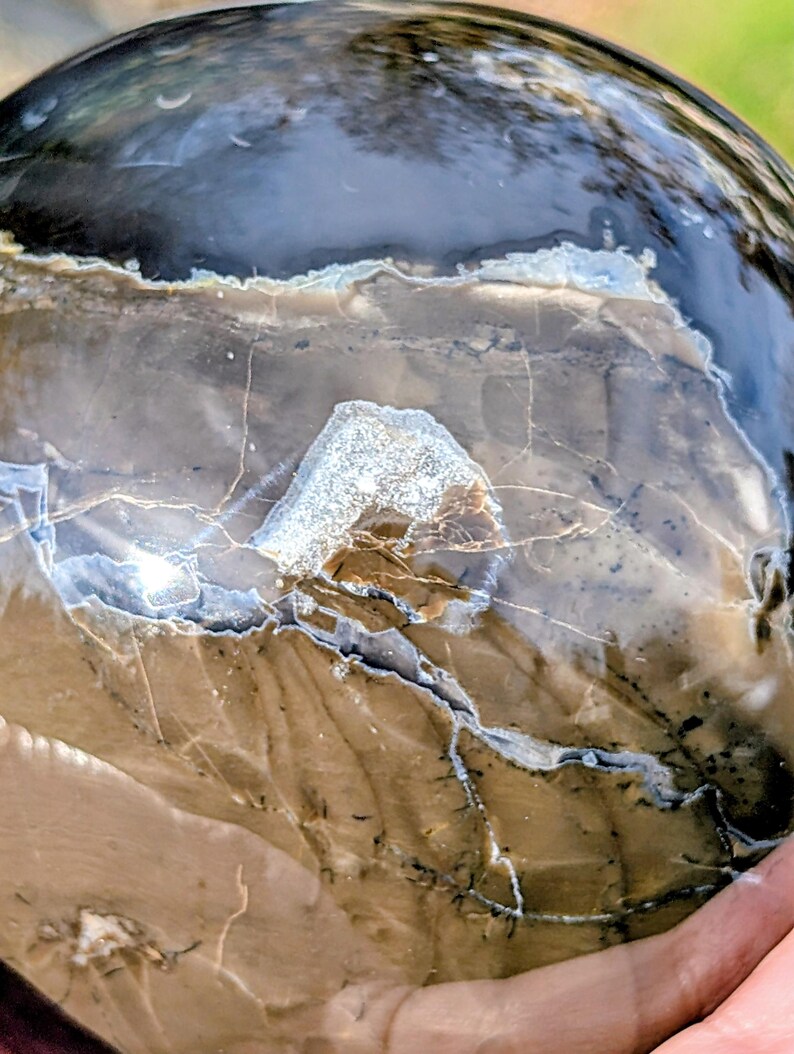 May include: A polished, spherical stone with a mix of brown, gray, and black hues. The surface features intricate patterns and veining, with a prominent, textured, light-colored area. The stone has a glossy finish, reflecting light.
